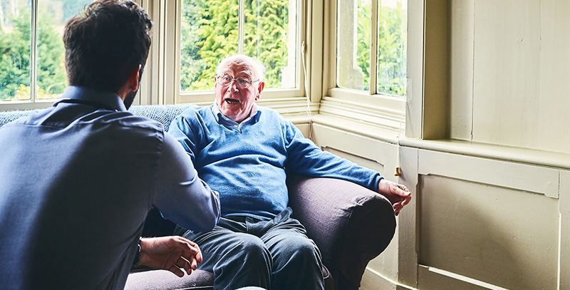 Older man talking while sat on couch in front of window