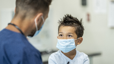 A young boy in a medical mask speaks with a healthcare professional
